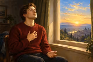 A young man in a crimson sweater prays beside a sunrise window with one hand over his heart and the other open on his lap.