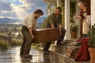 Two men share a heavy chest while a neighbor offers water after the rain.
