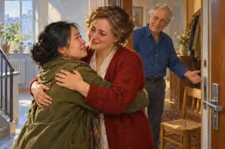 Two women embrace with relieved, tearful smiles at an apartment doorway while an older man welcomes them beside a wooden chair.